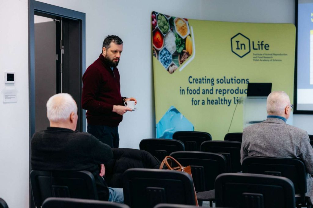 Mężczyzna z filiżanką w sali wykładowej na tle ścianki InLife PAN podczas seminarium naukowego.
