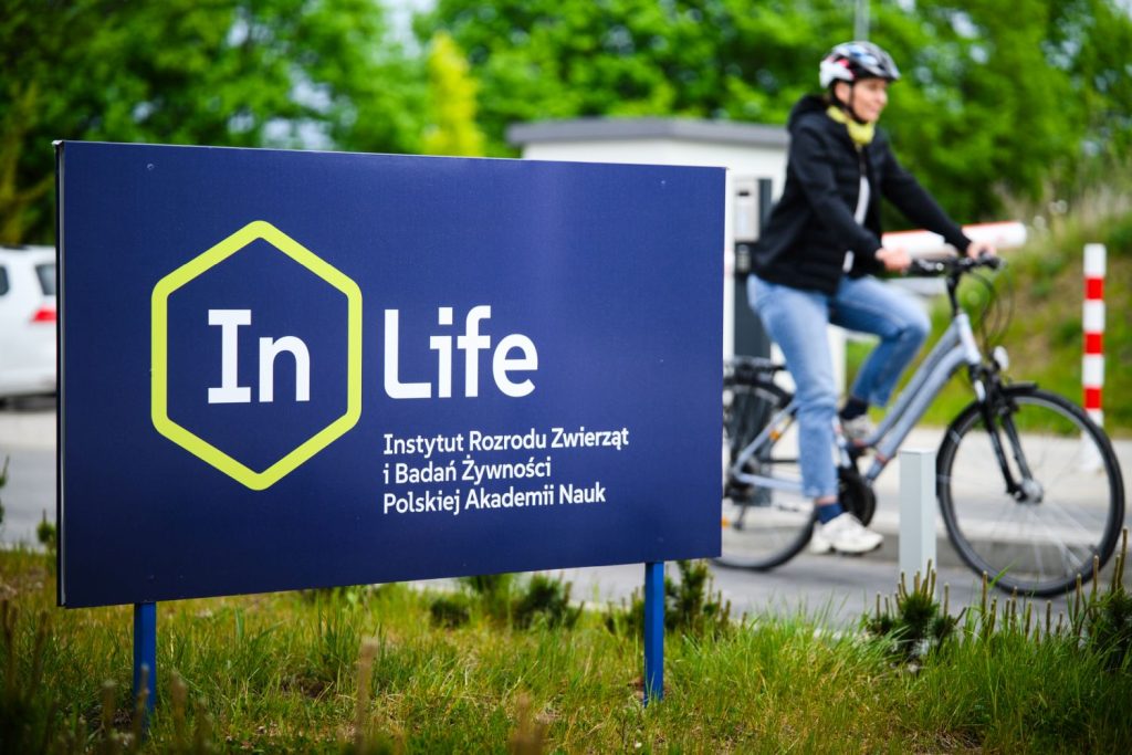 Rowerzystka mijająca tablicę Instytutu Rozrodu Zwierząt i Badań Żywności PAN – rozpoczęcie akcji Bike2InLife 2026.