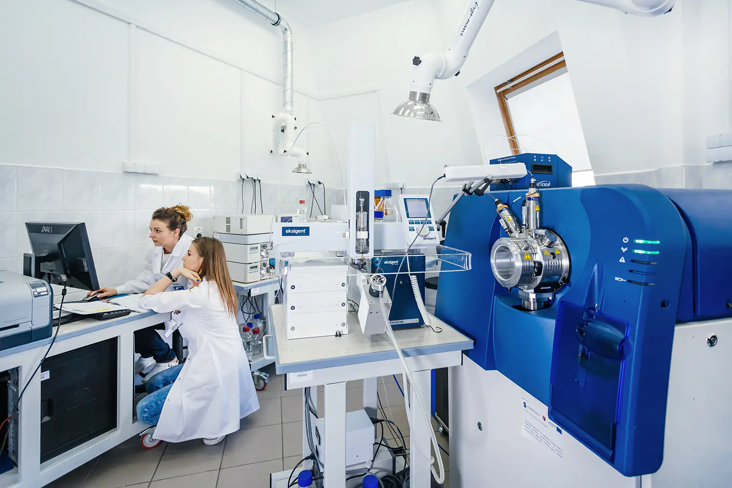 Dwie badaczki analizujące wyniki na komputerze obok nowoczesnego spektrometru mas w laboratorium.
