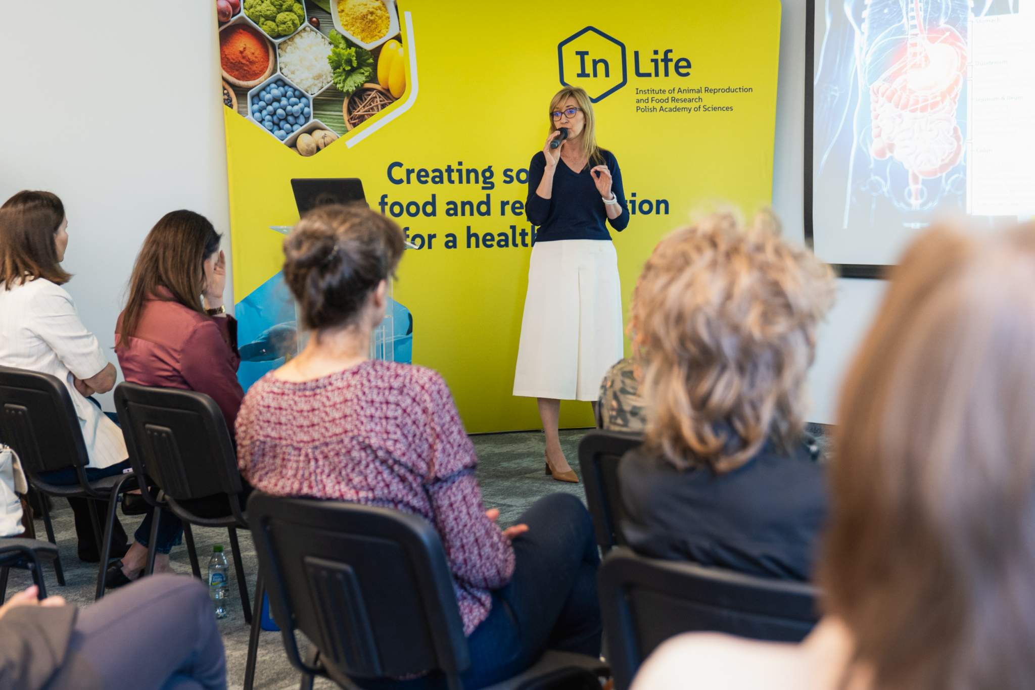 Prelegentka przemawiająca z mikrofonem na tle ścianki InLife PAN podczas seminarium naukowego.